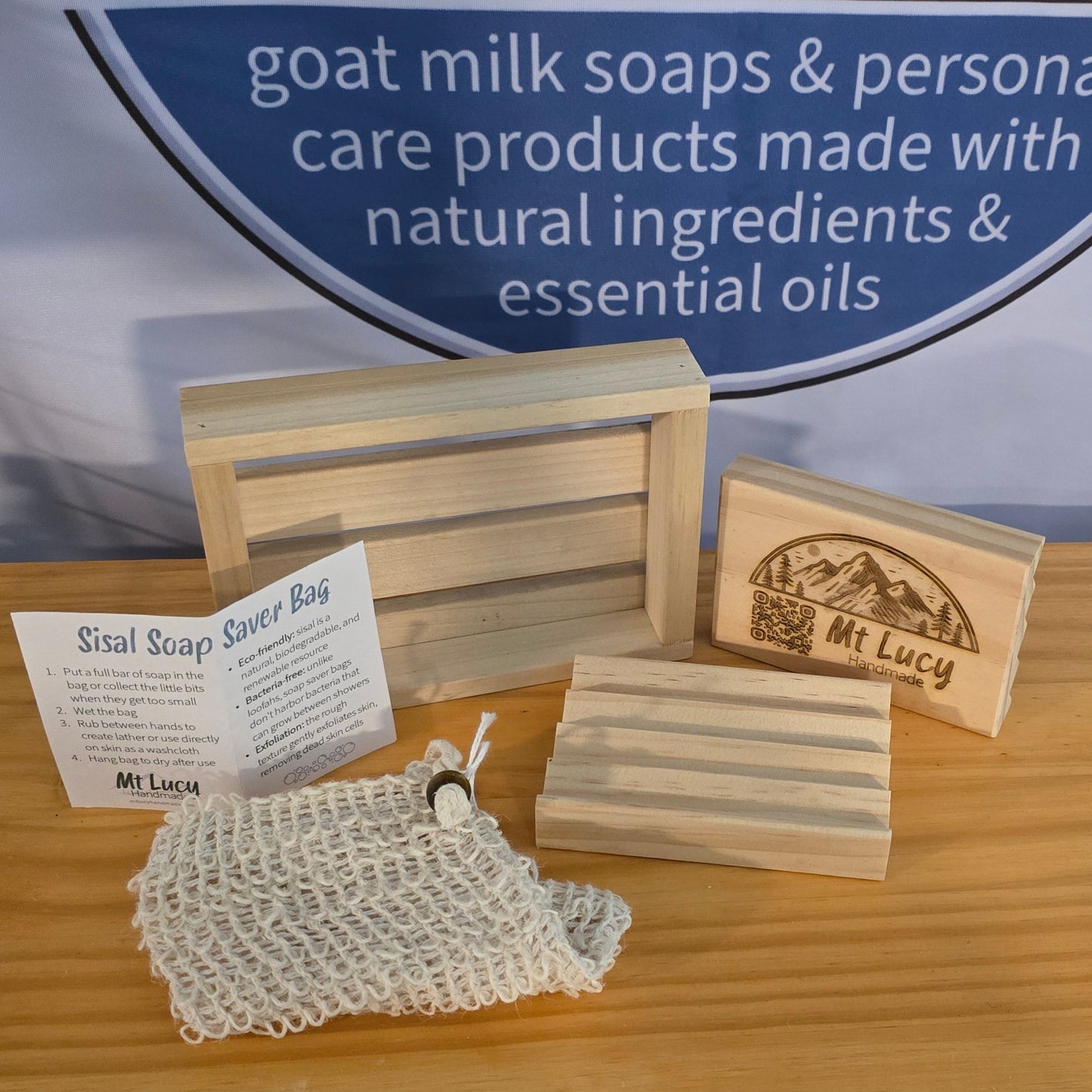 Soap dishes, soap crate, and soap saver bag on a wooden table with a branded backdrop.
