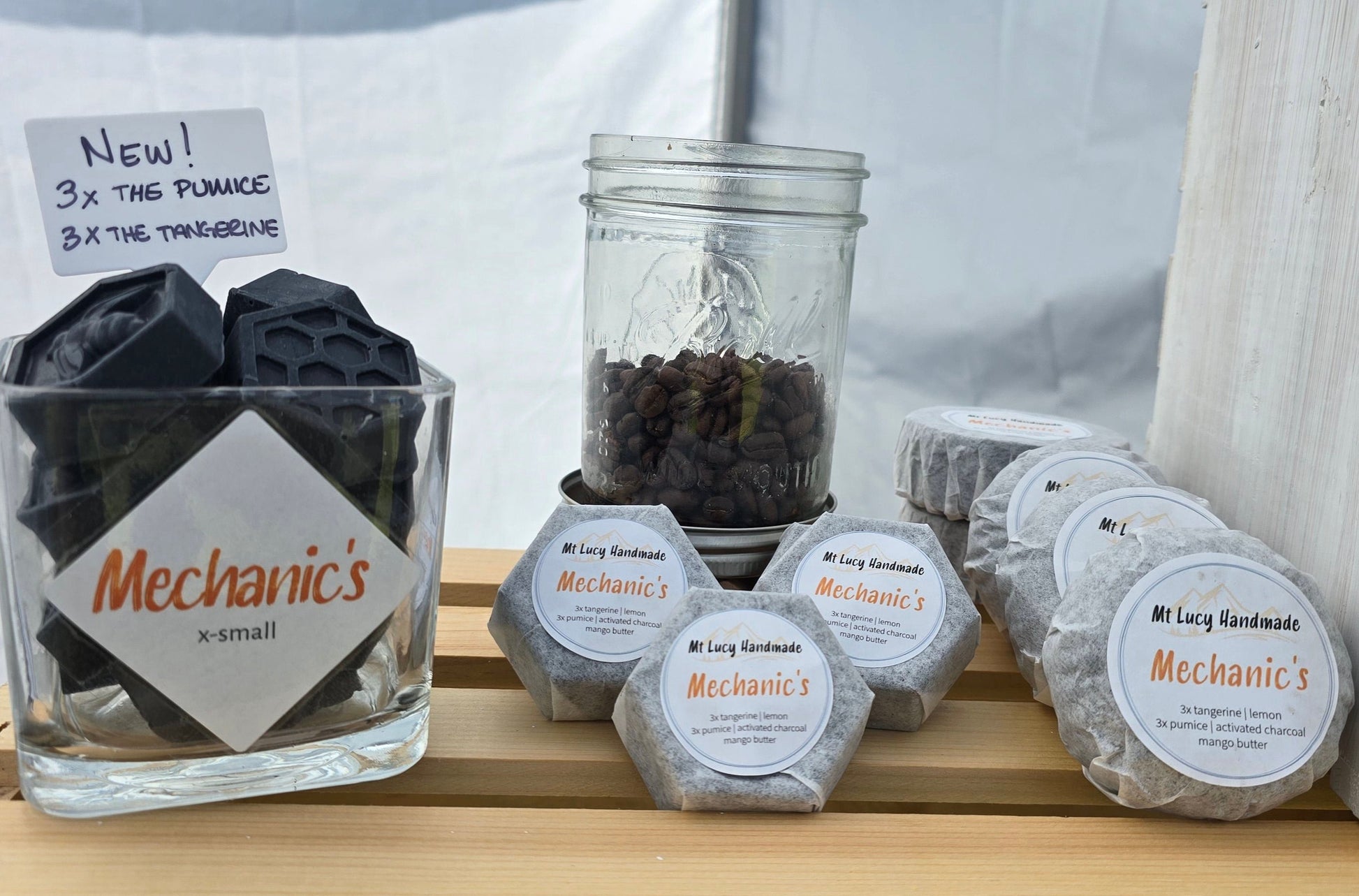 on the left, square glass jar filled with small hexagonal charcoal soaps - in the center, 3 packaged soaps in front of a jar of coffee - on the right larger packaged soaps in a line