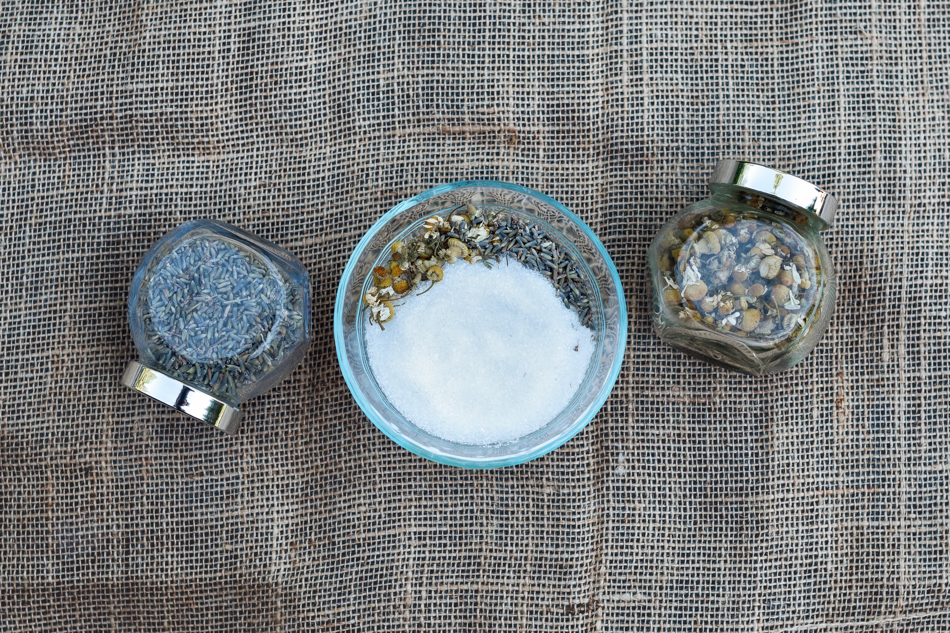 picture of a bowl of Epsom salts with lavender and chamomile on a burlap backdrop, with jars of lavender and chamomile on either side