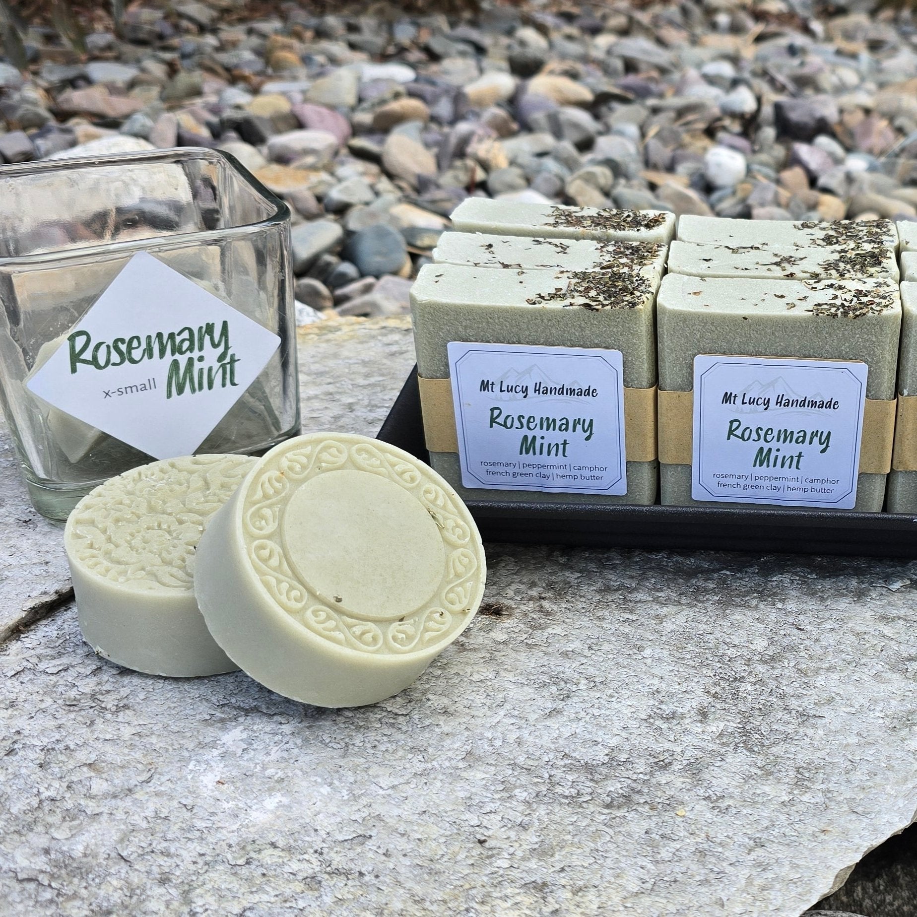 2 round, green-ish soaps on a flat stone slab, next to a black tray with 6 square bars of the same soap. 